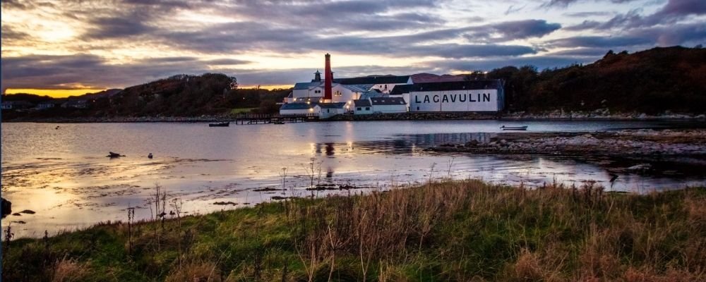 Lagavulin distillery on the shore of Islay at dusk, with its red chimney and whitewashed buildings reflected in the calm sea, a scene tied to the island's whisky festival, Fèis Ìle.