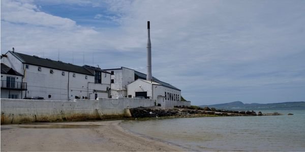 Bowmore distillery's whitewashed buildings and tall chimney beside Loch Indaal on Islay, with the sandy shore in the foreground during whisky festival season.