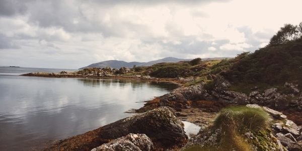 Rugged, rocky shoreline on Islay with calm sea, distant hills, and coastal scrub, near the island famed for its whisky festival, Fèis Ìle.